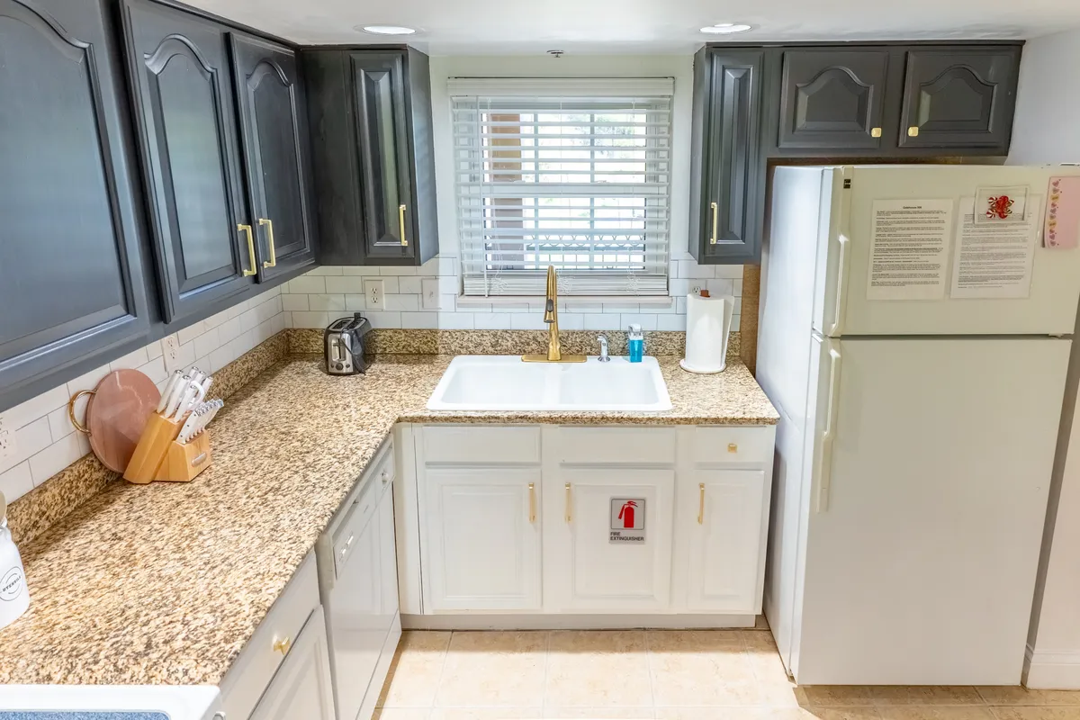 A warm and welcoming kitchen featuring a granite countertop, a kitchen sink, and elegant cabinets beneath a large window. The space is designed for cooking and family gatherings, with plenty of natural light and ample workspace for meal preparation.