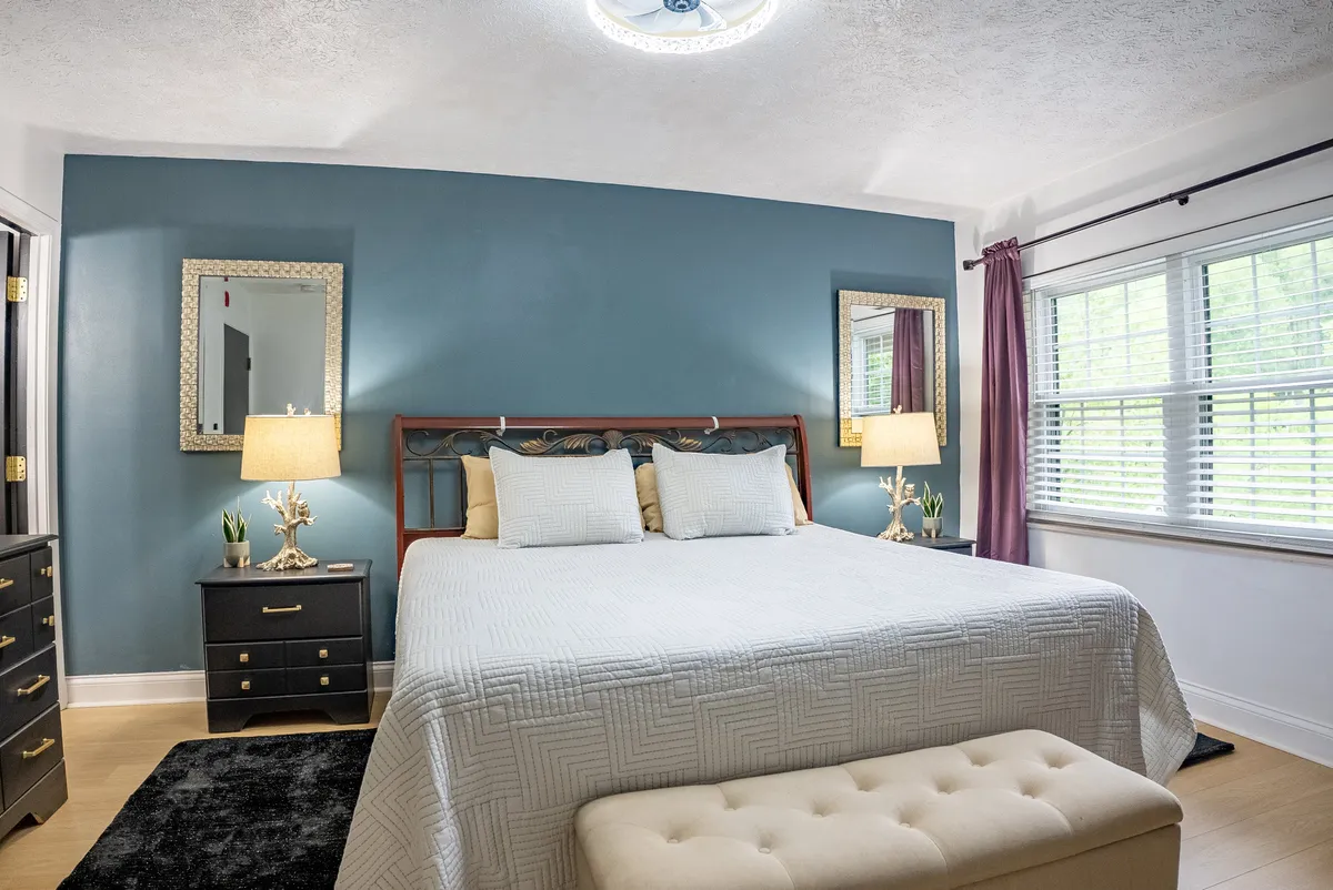 This image features a bed with a white quilt set against a blue accent wall, flanked by mirrors and softly glowing table lamps. Natural light filters into the room, creating a tranquil and cozy atmosphere ideal for relaxation and unwinding.