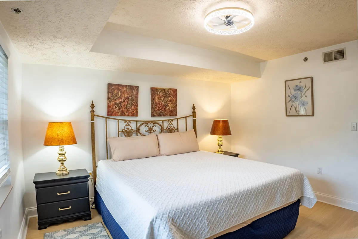 A tranquil bedroom setup with a bed, bedside tables, lamps, and artwork. The soft lighting and neutral color palette evoke a sense of peace and comfort, suggesting a welcoming place for relaxation and quiet mornings.
