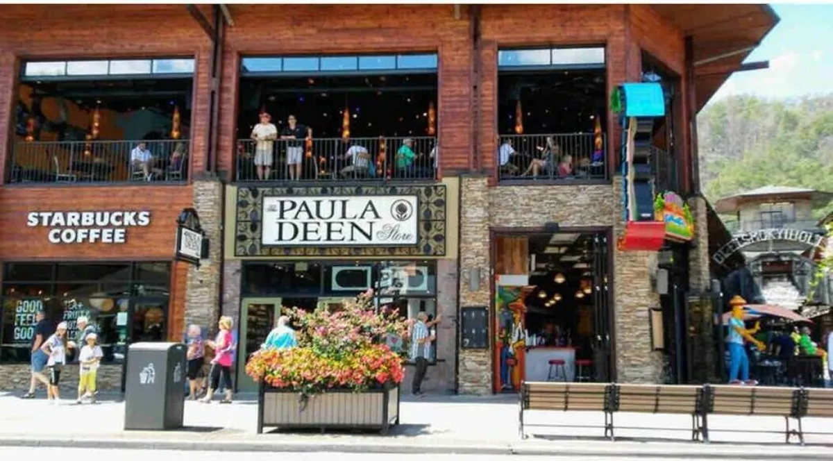 A lively downtown street scene featuring the Paula Deen Store and Starbucks Coffee signs, vibrant flowers, and people socializing on balconies. The view captures the energetic and welcoming atmosphere of Gatlinburg’s local dining and shopping scene.