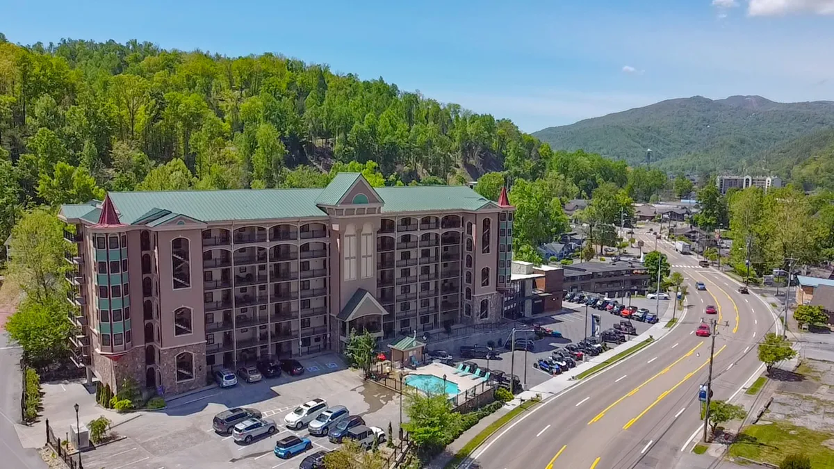 The image showcases the exterior of a modern condo building in Gatlinburg surrounded by lush greenery, with a sparkling outdoor pool as the central feature. Guests can be seen lounging by the pool and enjoying the relaxed atmosphere. The setting sun casts a warm glow over the scene, suggesting the appeal of sunset views from the balconies.