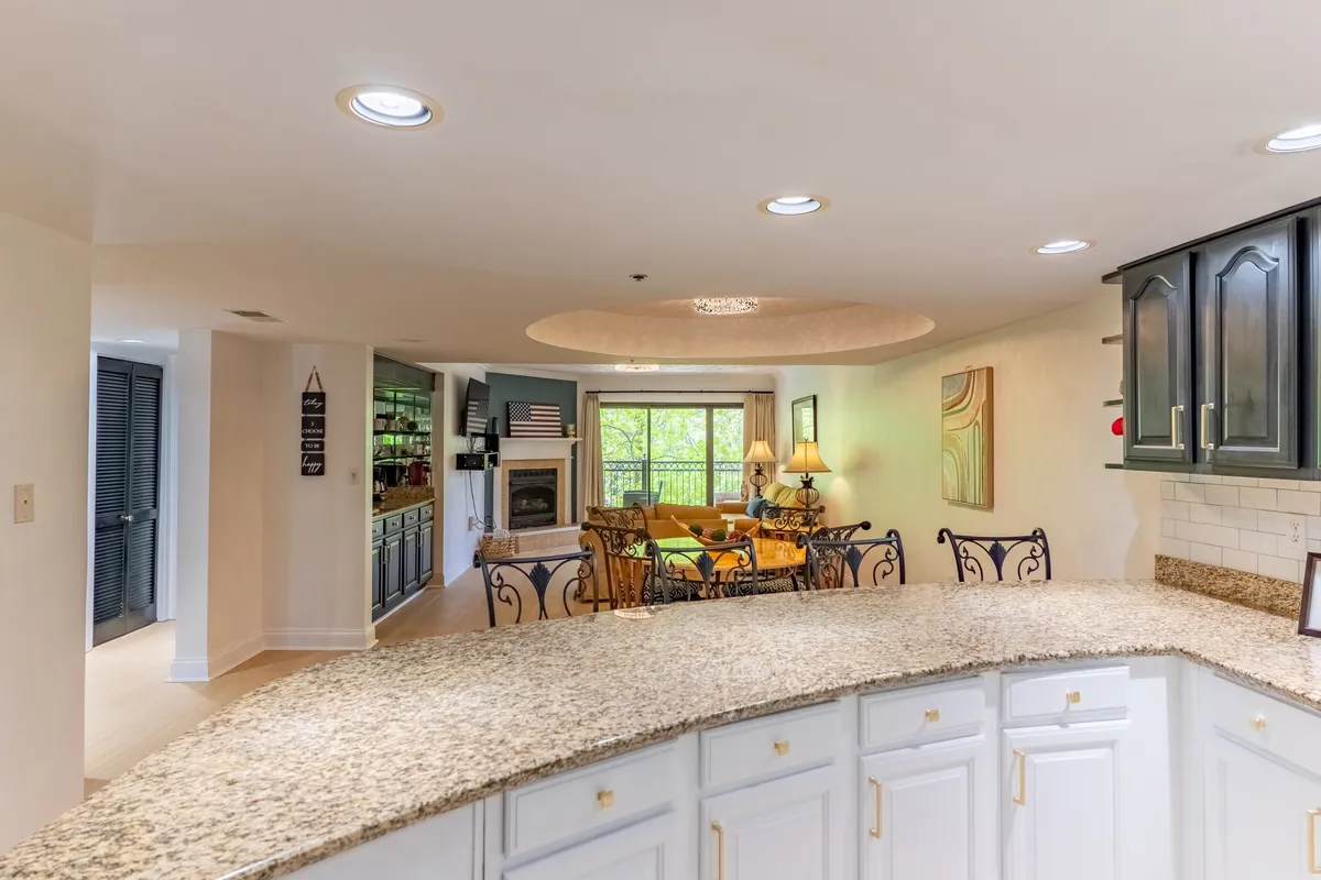 A modern, welcoming open-plan kitchen and living area with a granite countertop, stylish bar stools, and a fireplace. The living area features bright, cheerful furniture, while the kitchen space invites easy entertaining and social interaction. The fireplace anchors the room, providing a cozy focal point for gatherings.