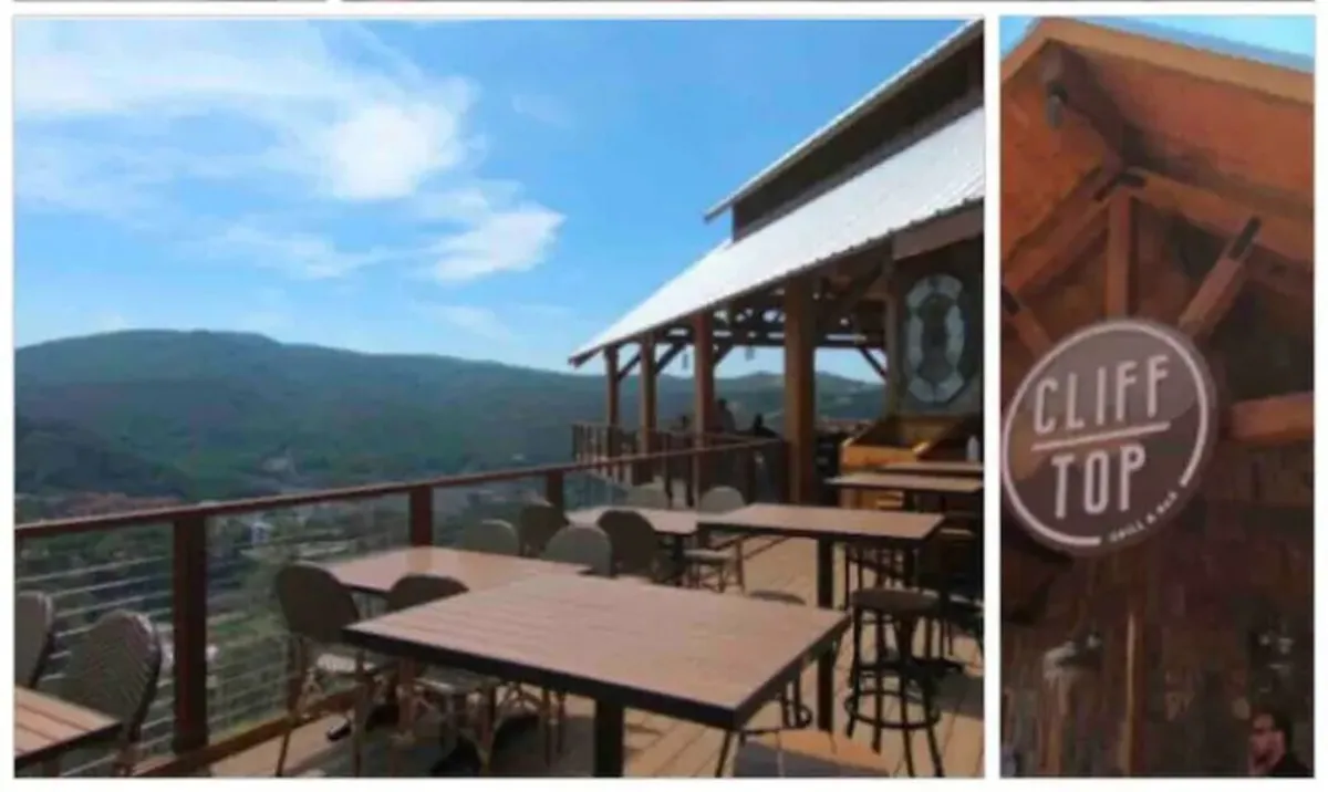 An outdoor restaurant seating area with tables and chairs set against a backdrop of scenic mountain views. The setting sun bathes the space in warm light, with the restaurant signage visible and lush greenery surrounding the perimeter, creating a sense of relaxation and romance.