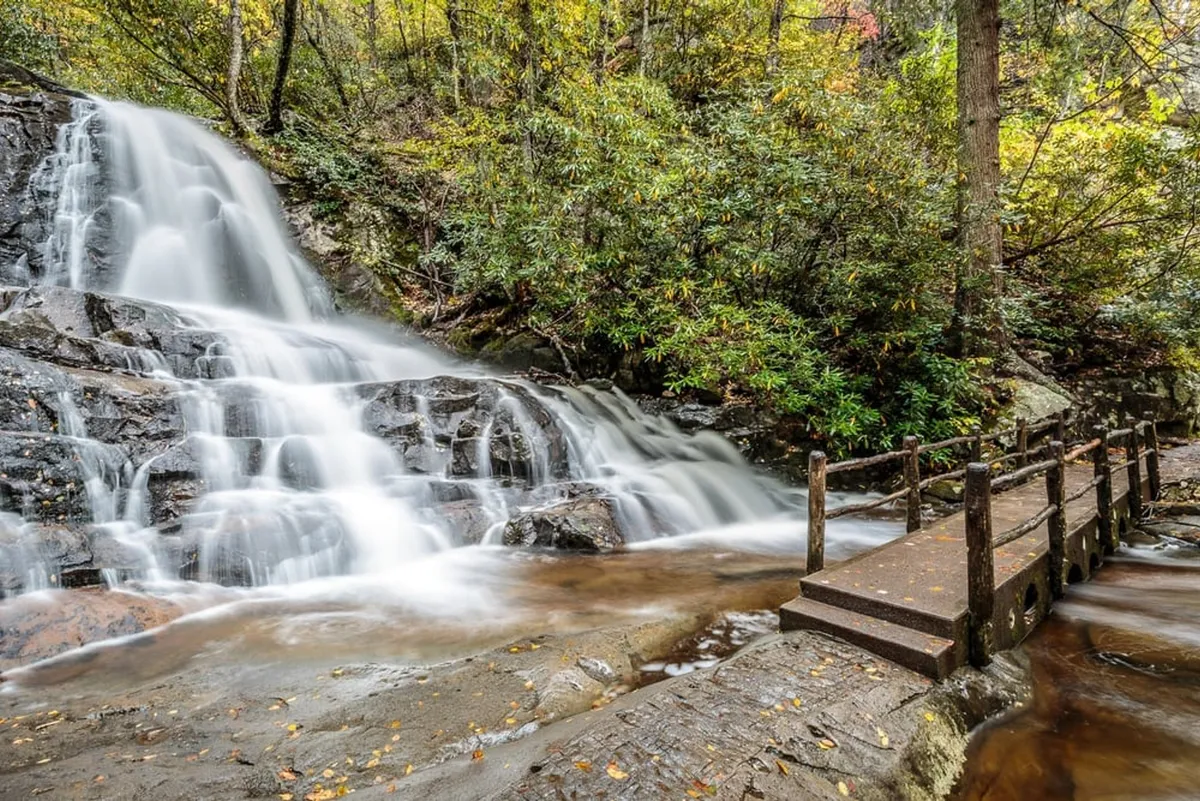 A picturesque waterfall cascades beside a wooden bridge surrounded by lush green trees, capturing a classic Smokies scene. The tranquil water and natural setting evoke the allure of nearby hiking trails and waterfall destinations.
