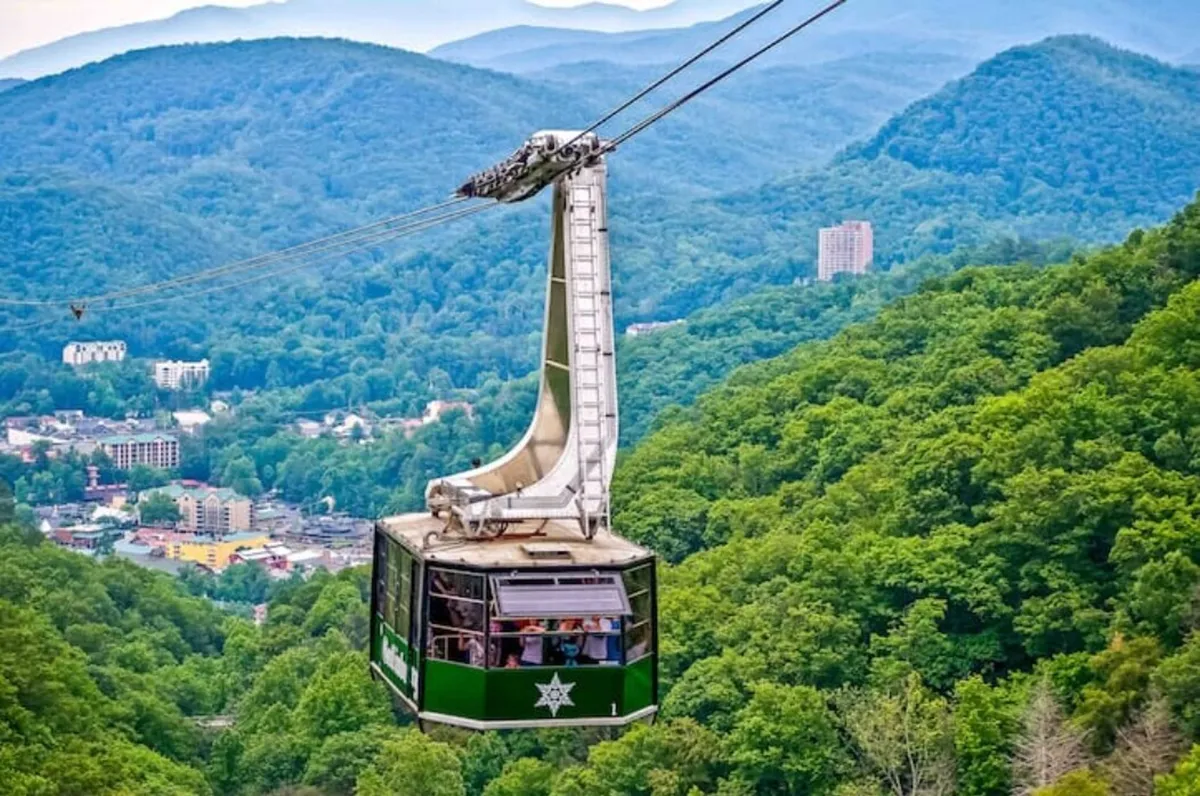 A dramatic panoramic shot showcasing a gondola ascending above Gatlinburg, framed by a sweeping view of the Smoky Mountains, green trees, and the town below. The image suggests adventure and the thrill of exploring local attractions like Anakeesta.